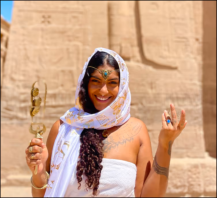 Smiling woman with curly hair wearing white traditional attire and jewelry, standing in front of ancient stone carvings.