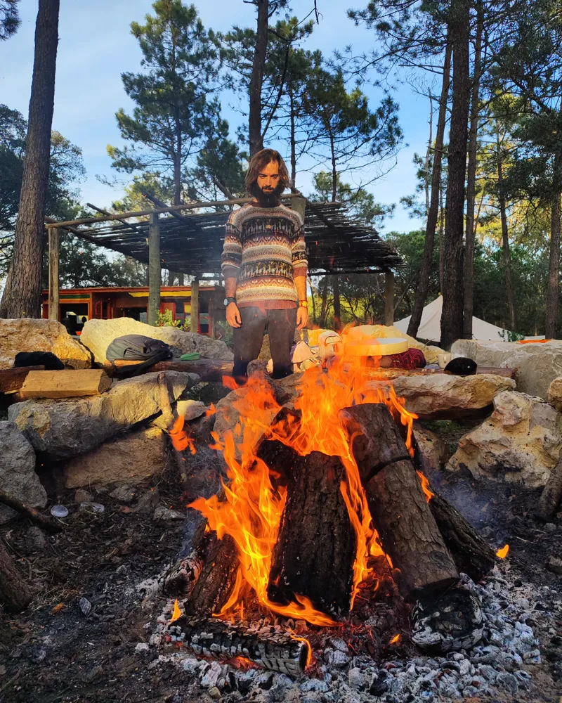 Tiago standing behind a fire he keeps, among stones and pine trees.