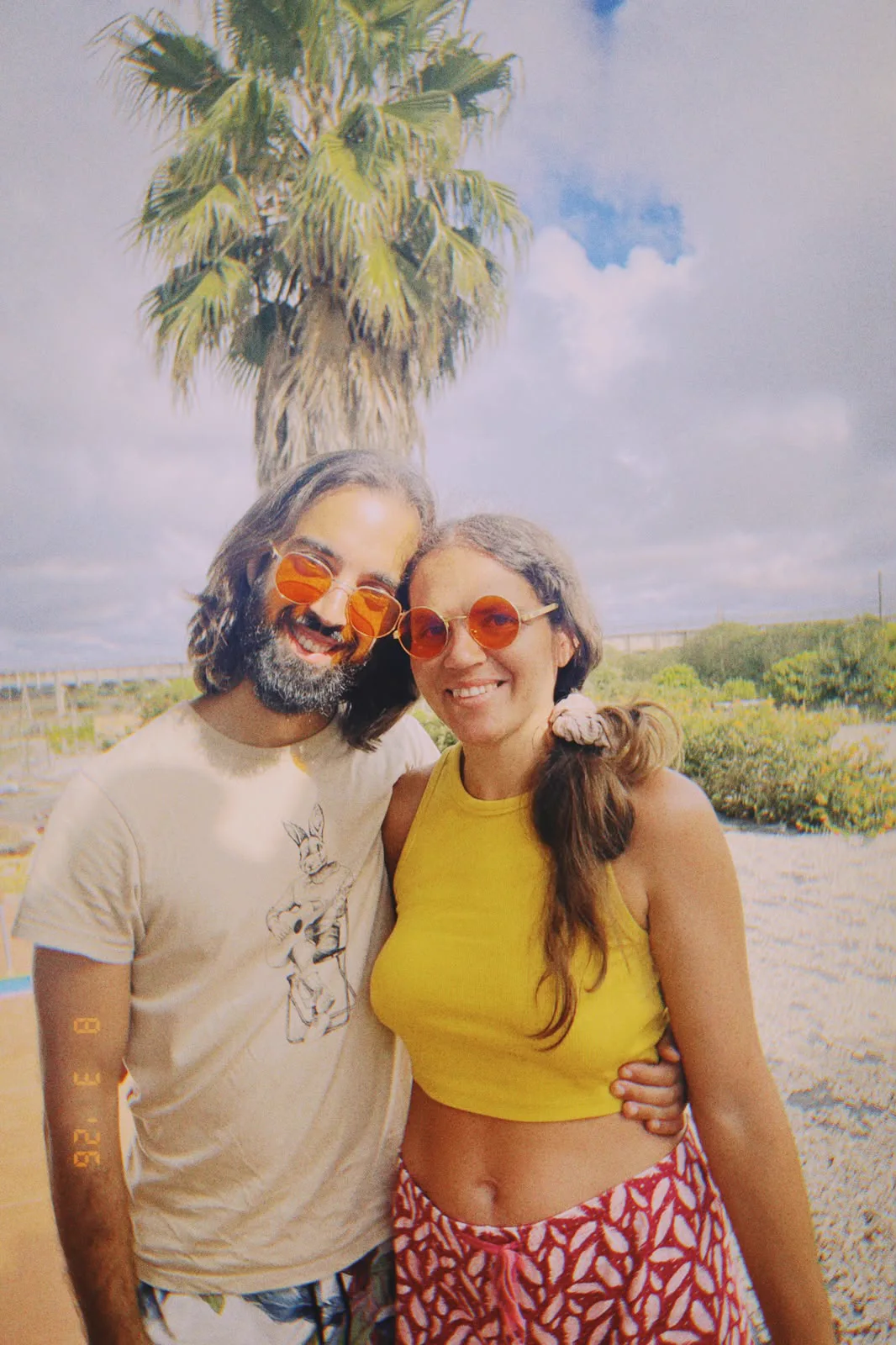 Tiago and Irina standing close in the sun, both in round orange sunglasses, a palm tree behind them.
