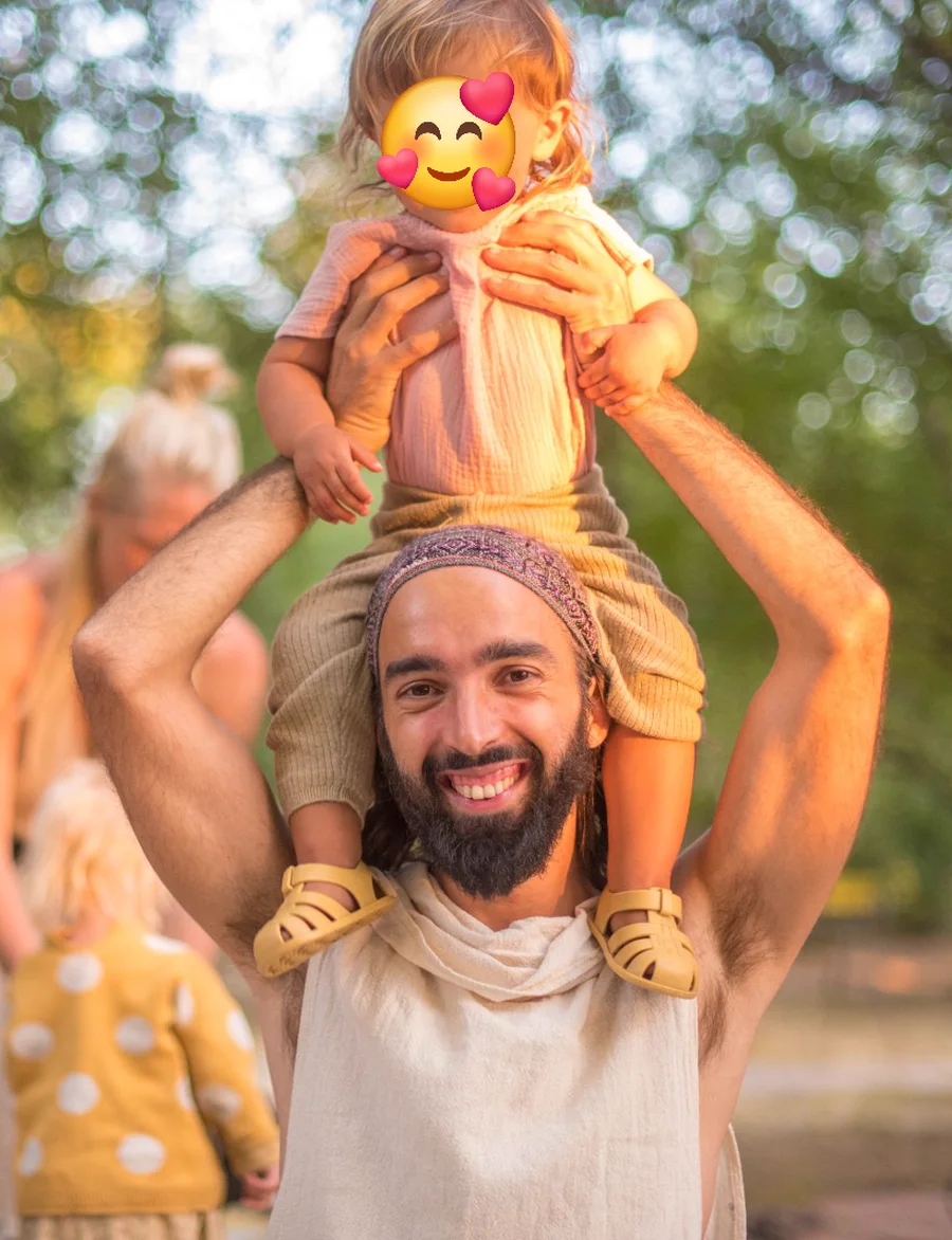 Tiago smiling with his son on his shoulders, among trees in the late afternoon light.