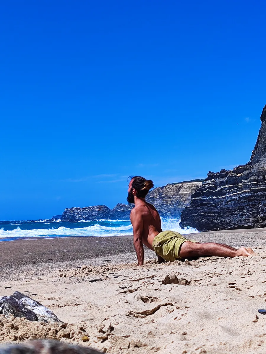 Tiago in a cobra pose on an empty beach, waves breaking against dark cliffs behind him.