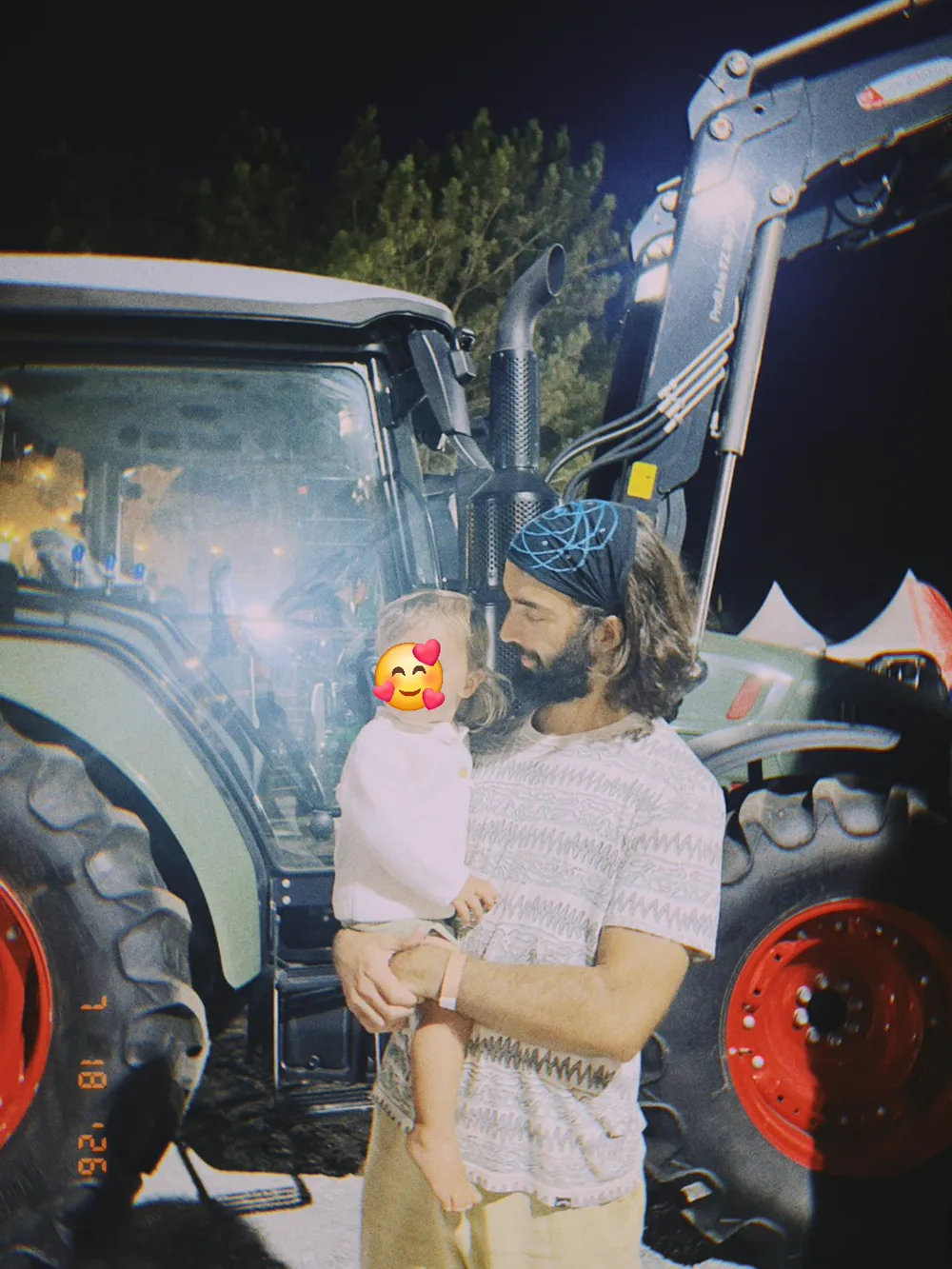 O Tiago à noite em frente a um trator verde grande de rodas vermelhas, com o filho ao colo, a cara dele tapada por um emoji