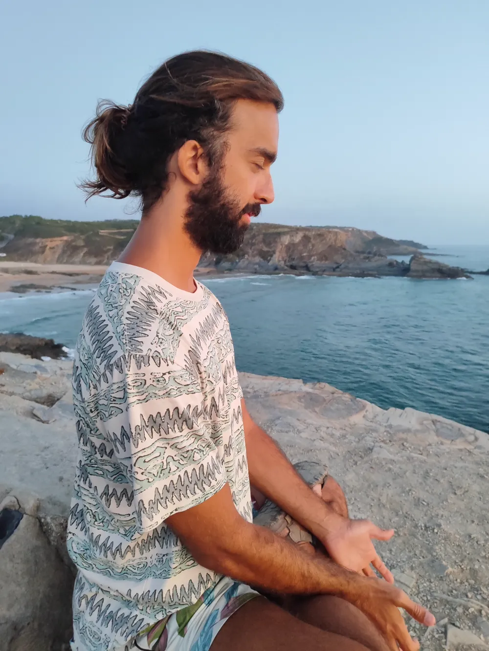 Tiago sitting cross-legged on a rock above the sea at dusk, eyes lowered, hands open on his knees