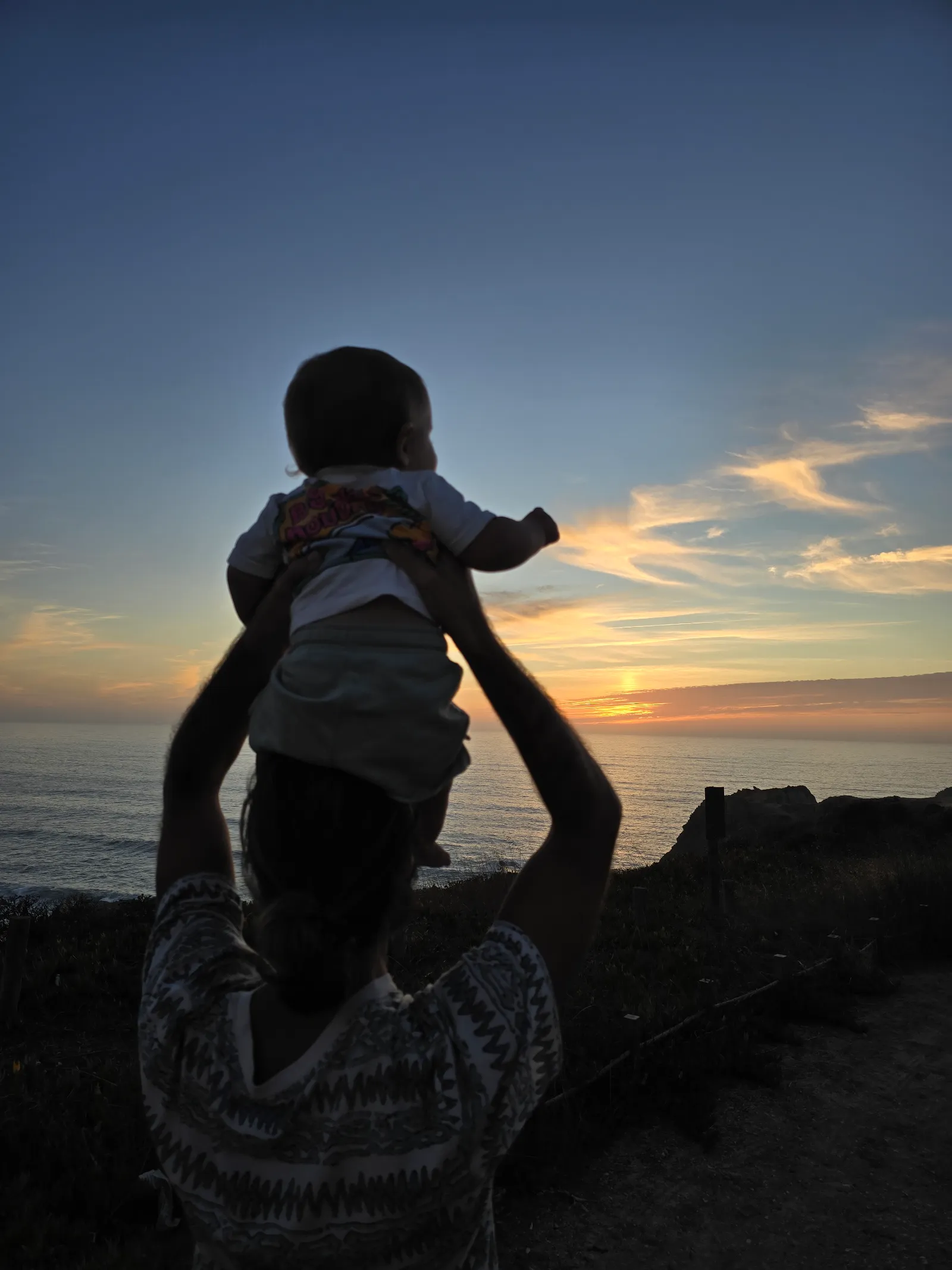 Tiago from behind, holding his son up against a sunset over the sea