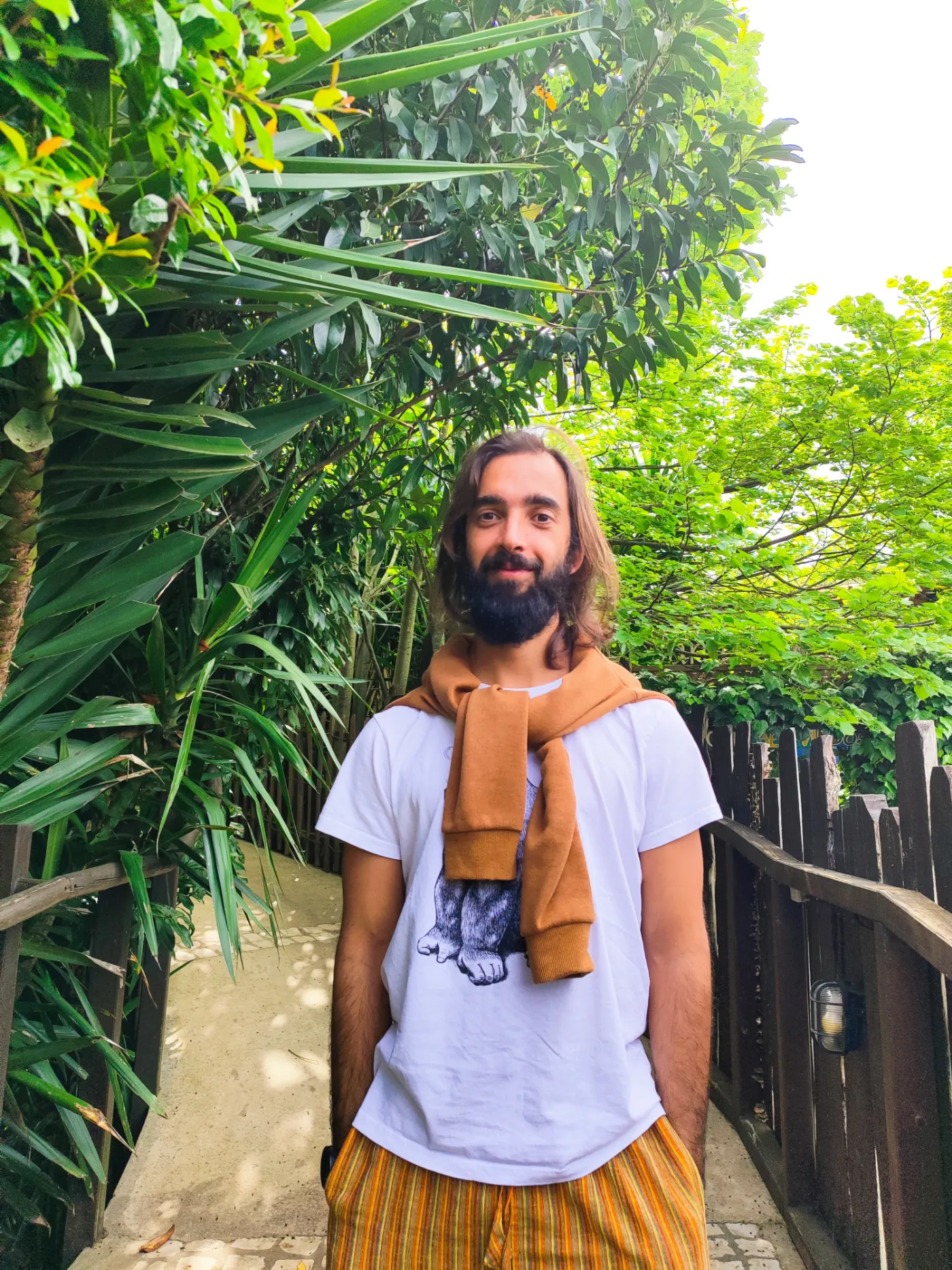 Tiago smiling at the camera between tall green plants, with an orange jumper over his shoulders