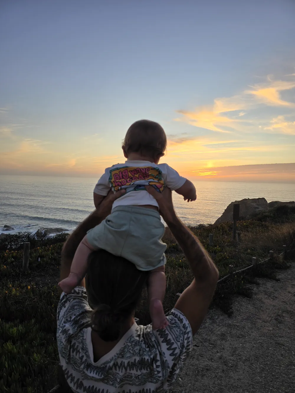 O Tiago de costas ao pôr do sol sobre o mar, a levantar o filho para os ombros