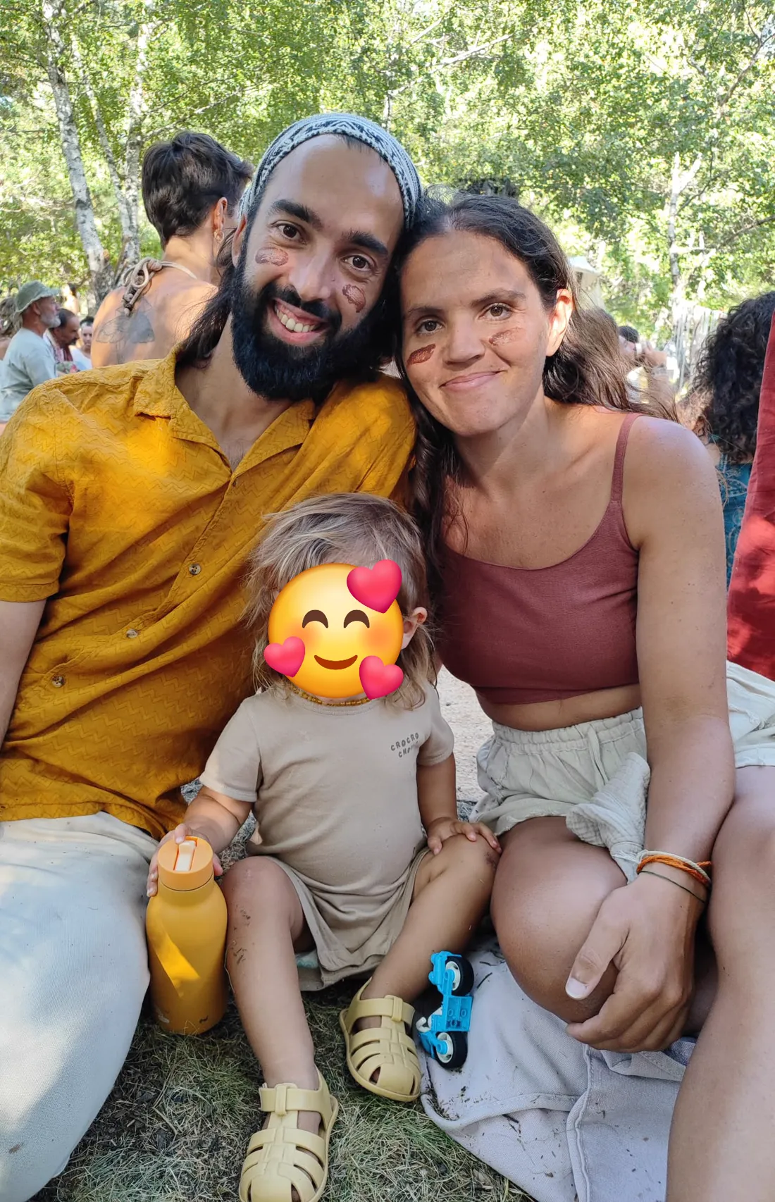 O Tiago e a Irina sentados na relva num encontro, com o filho entre os dois, a cara dele tapada por um emoji