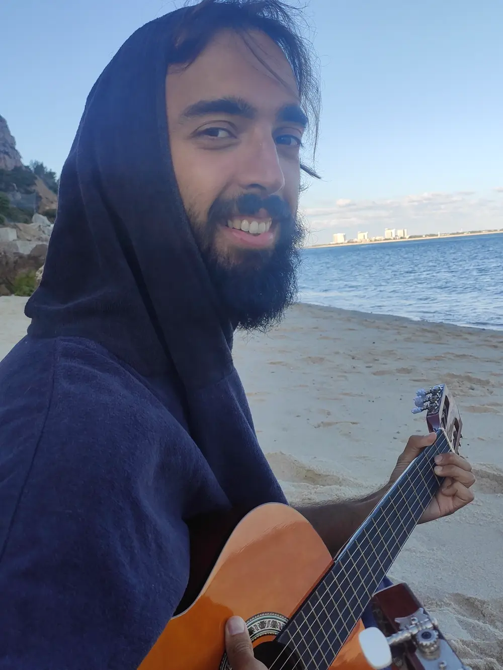 O Tiago de capuz numa praia, a sorrir para a câmara, com uma guitarra
