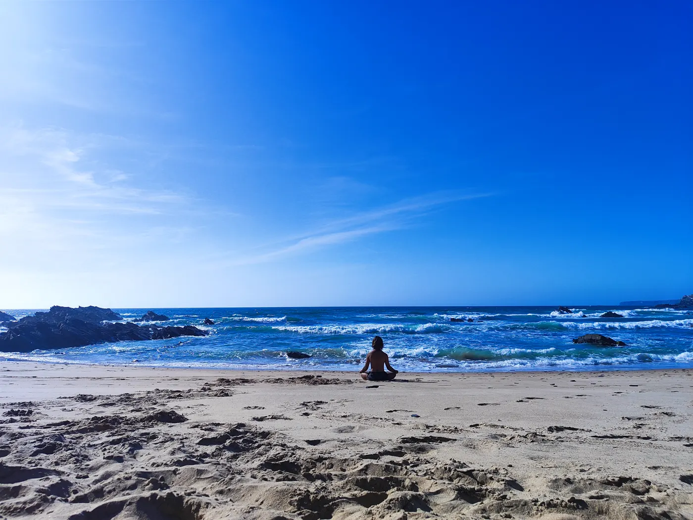 Tiago meditating, very small, on a wide empty beach, with the sea and the rocks behind him