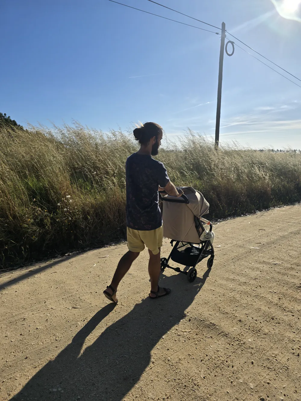 O Tiago de costas numa estrada de terra no Alentejo, a empurrar um carrinho de bebé entre erva alta e seca, debaixo de um céu azul
