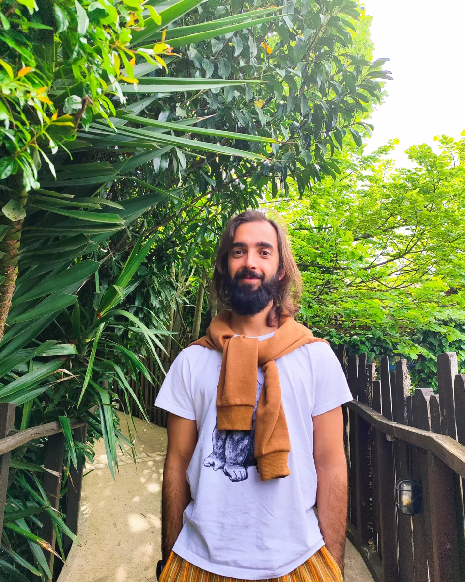 Tiago smiling at the camera among tall plants, a jumper tied over his shoulders.