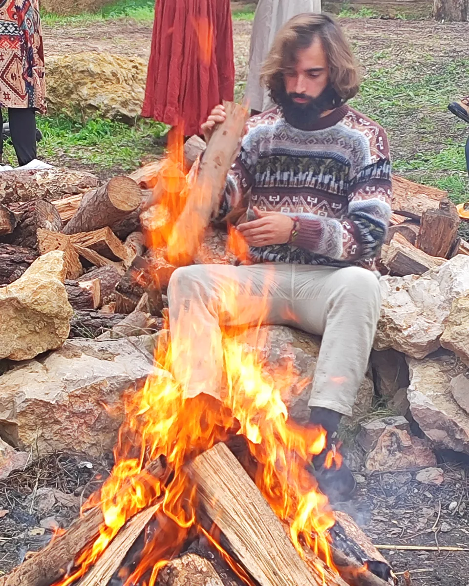 Tiago feeding a log into a big outdoor fire, in a wool jumper.