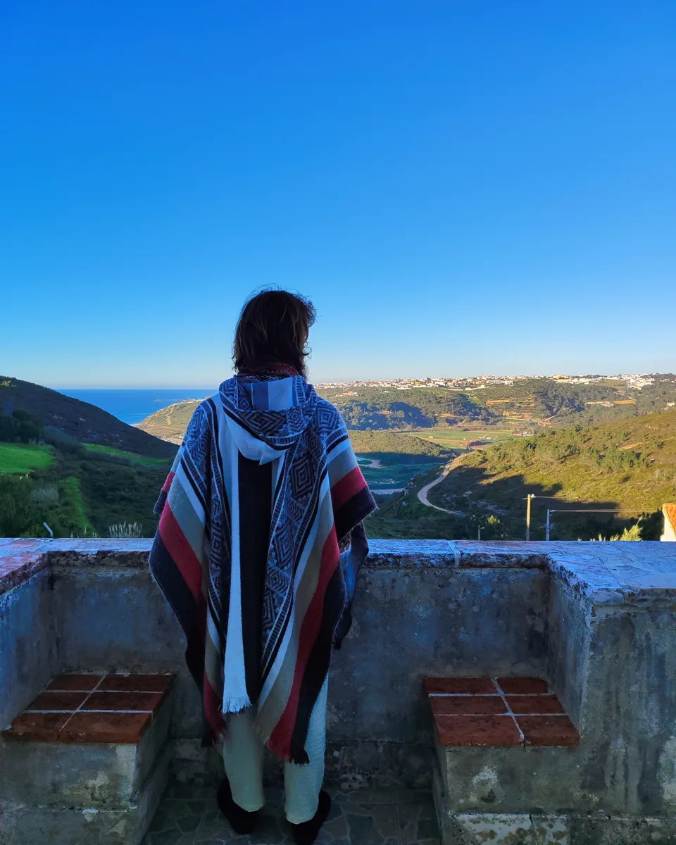Tiago seen from behind in a striped poncho, looking out over a green valley towards the sea.