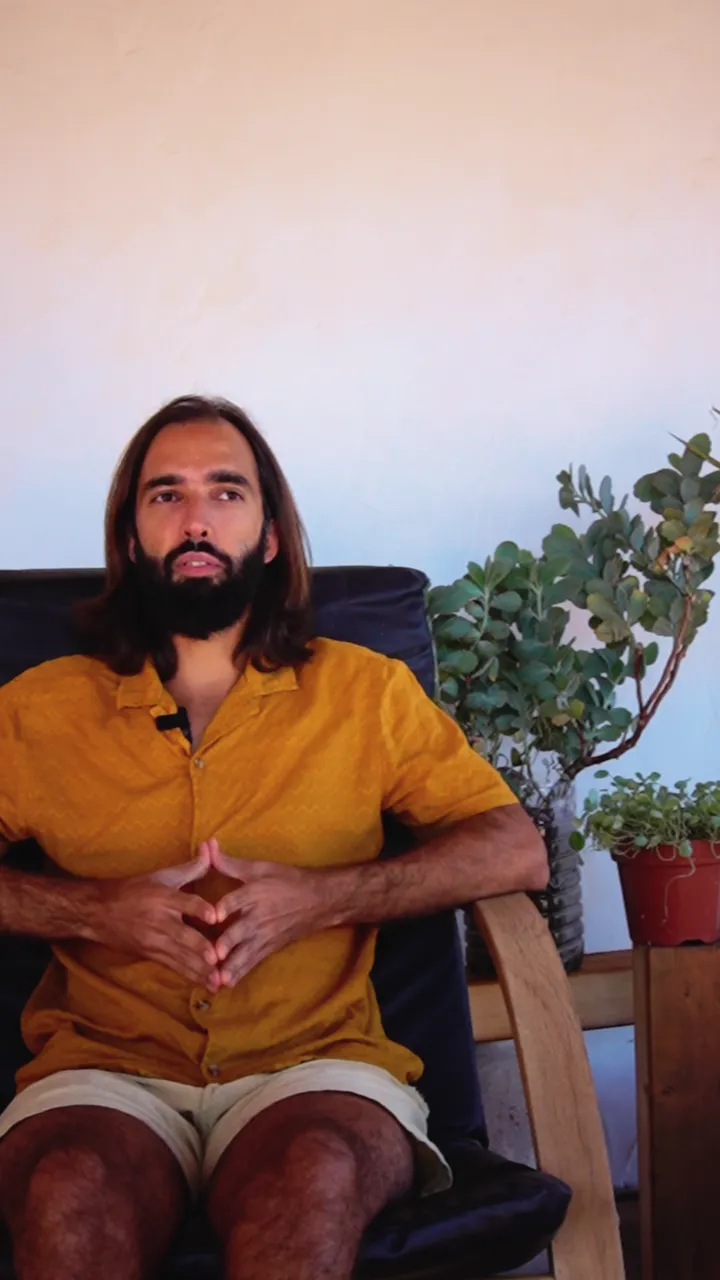 O Tiago de camisa amarela, sentado numa cadeira de madeira num terraço com plantas, a falar para a câmara